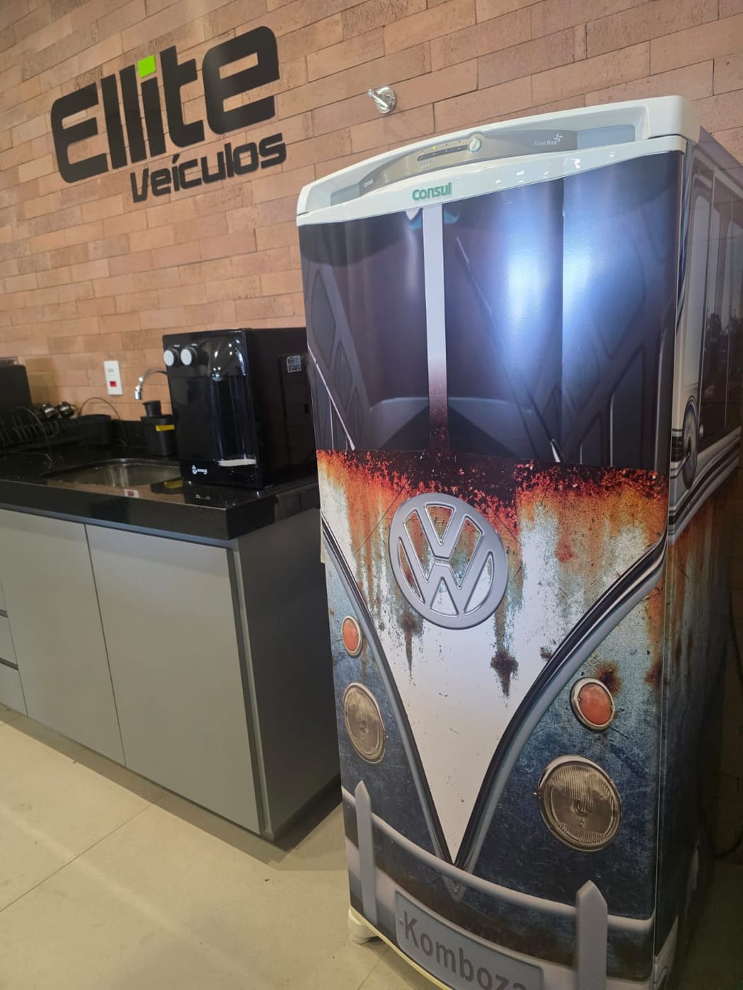Copa da loja com a geladeira envelopada como a frente de uma Kombi antiga, ao lado da máquina de café e do logotipo Ellite Veículos na parede de tijolos